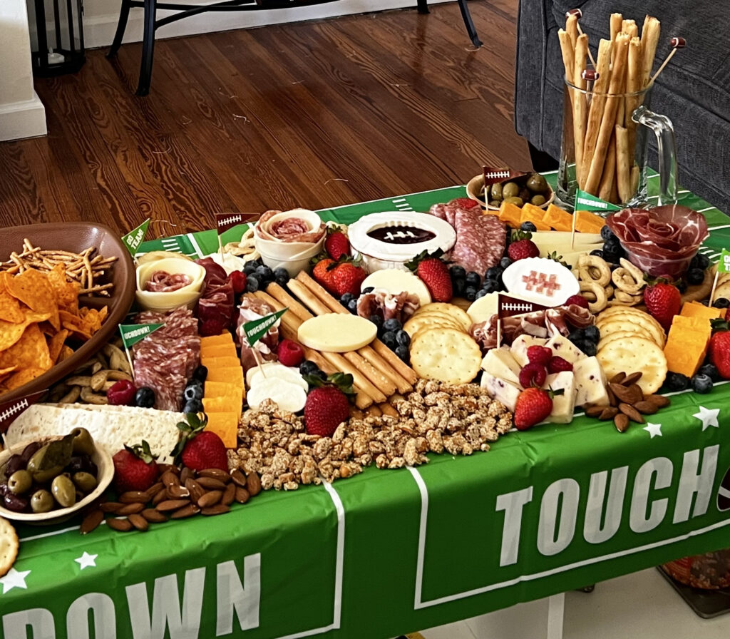 A large, football-themed grazing table, loaded with fresh, cheese, charcuterie, produce, and breads